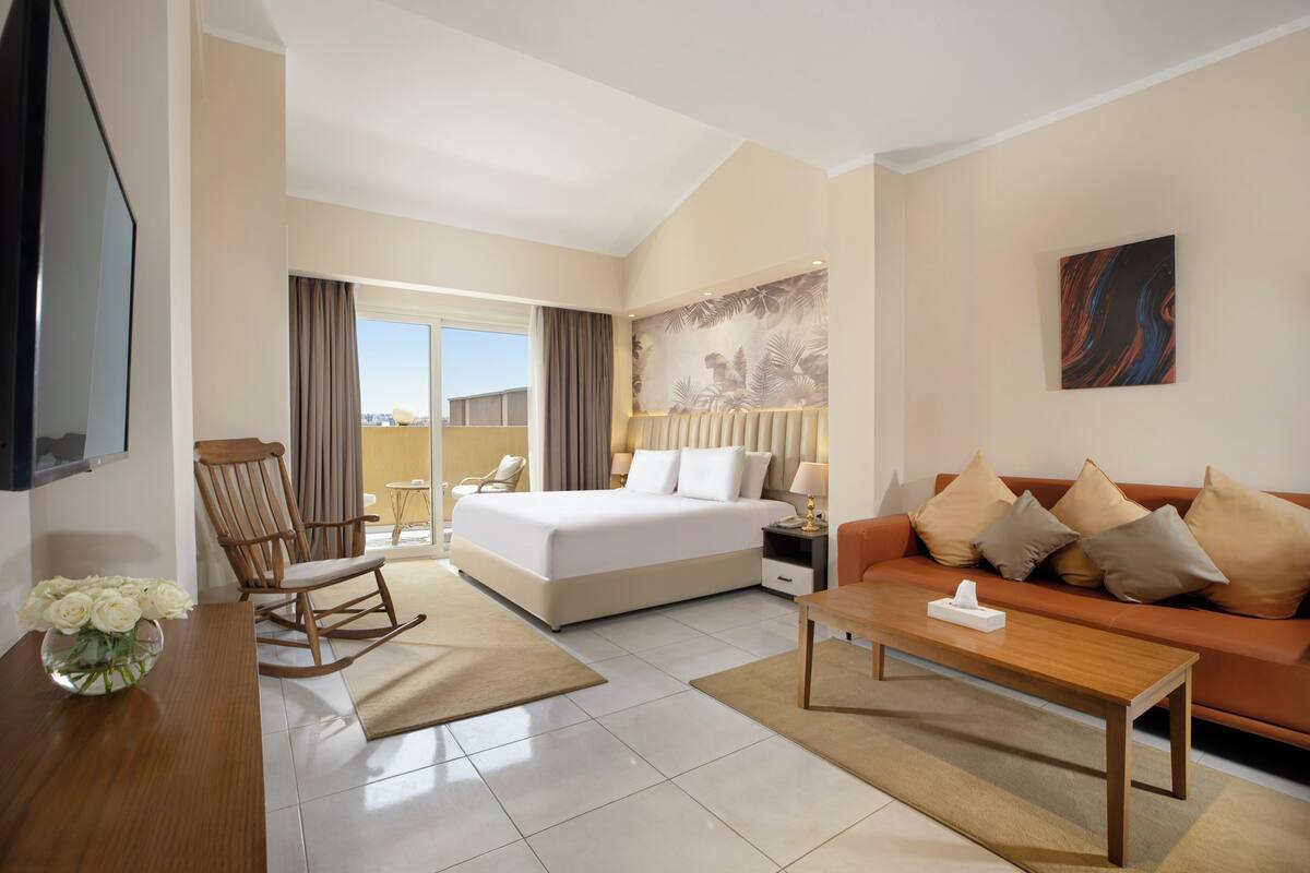 A hotel room with a bed, a sofa, a coffee table, a rocking chair, a TV, and a painting on the wall.