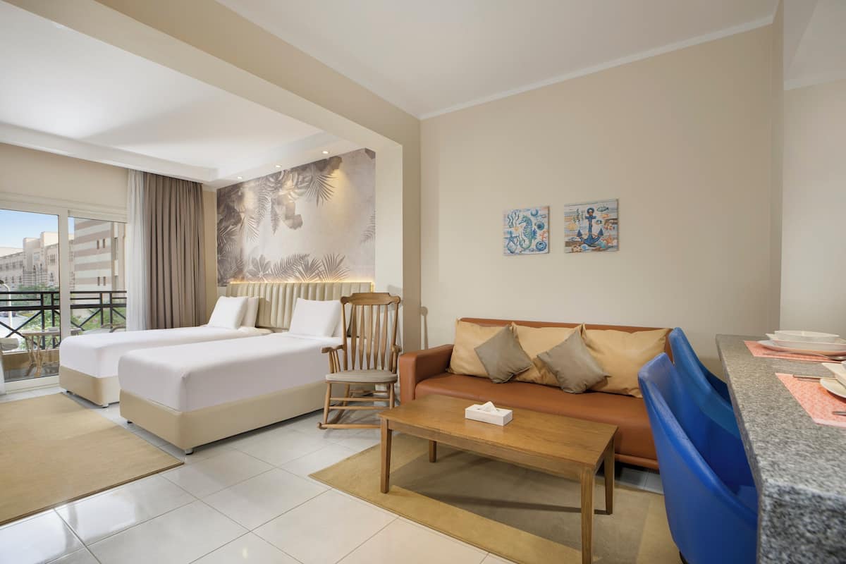 A hotel room with two beds, a sofa, a wooden coffee table, and a balcony with a view of buildings.