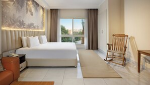 A modern hotel room with a large bed, a wooden rocking chair, a small table, and a view of the city through large windows. - Vibotel Residence Hotel Apartments (New Cairo)
