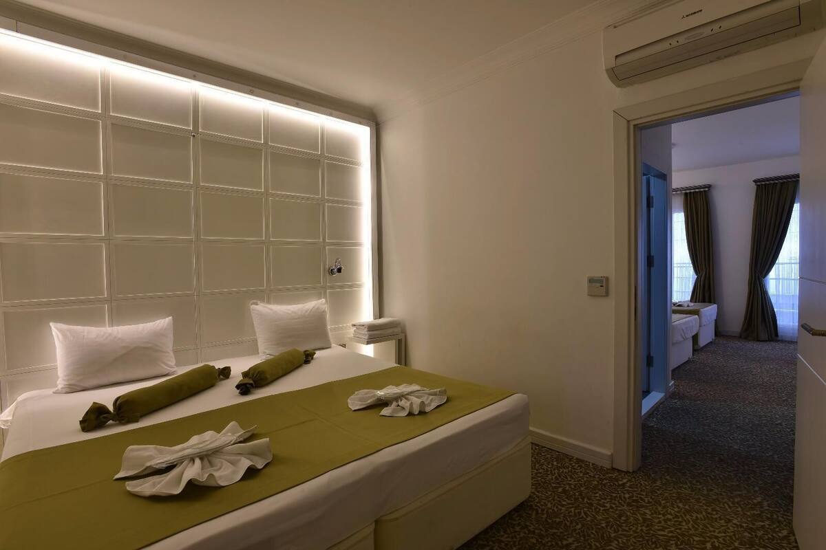 A hotel room with a large bed, a bedside table, a wall-mounted air conditioner, and a view of another room through an open door.