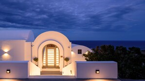 A white building with an arched entrance, illuminated by exterior lights, and a view of the sea in the background. - LF Suites & Villas (Santorini)