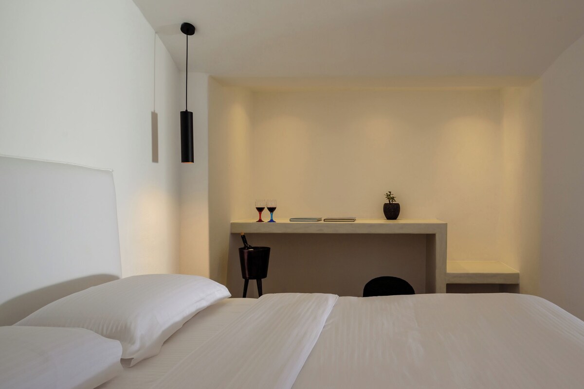 A minimalist hotel room with a large bed, a small desk with a plant, and a chair.