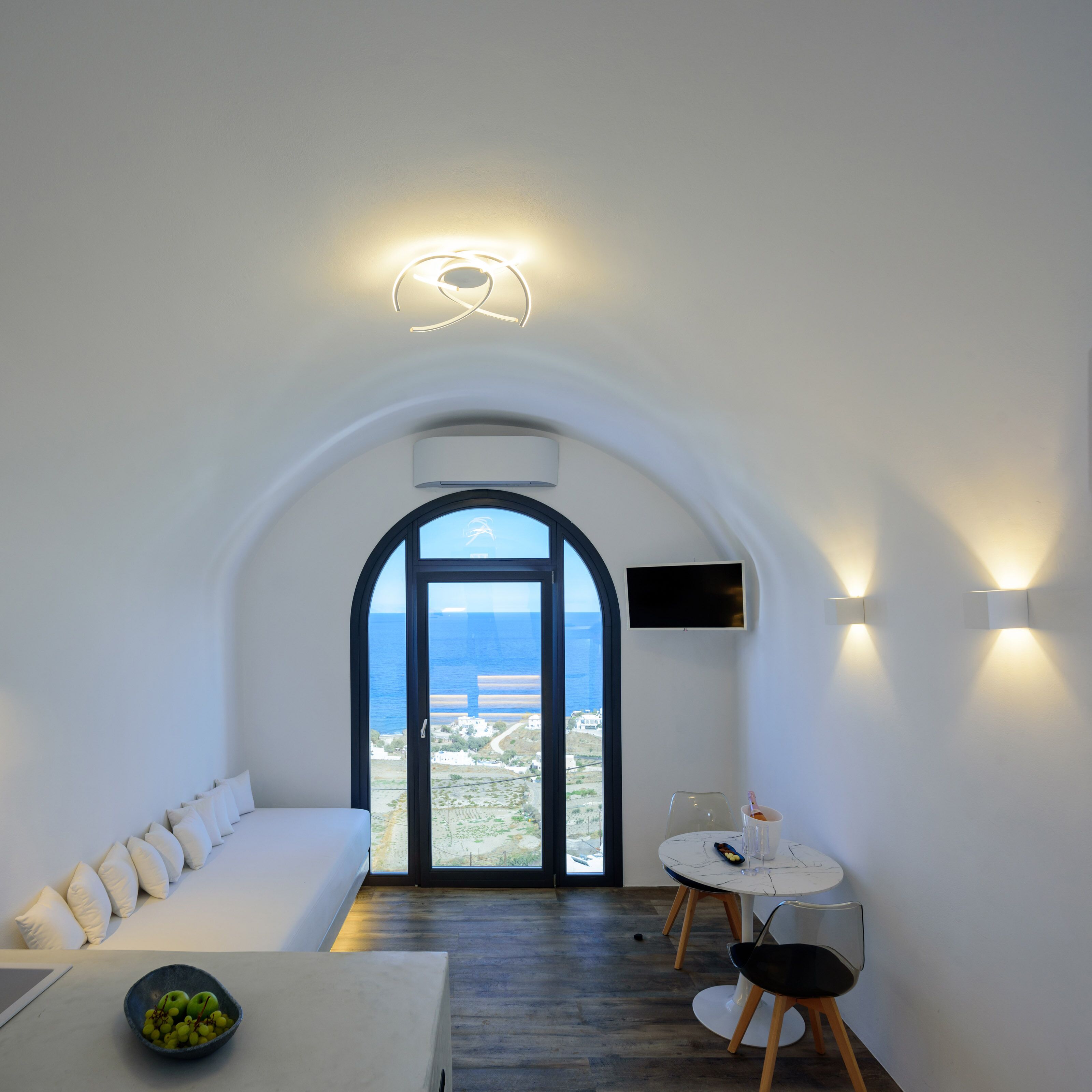 A modern room with a white couch, a dining table with chairs, a bowl of fruit, and a view of the sea through an arched doorway.