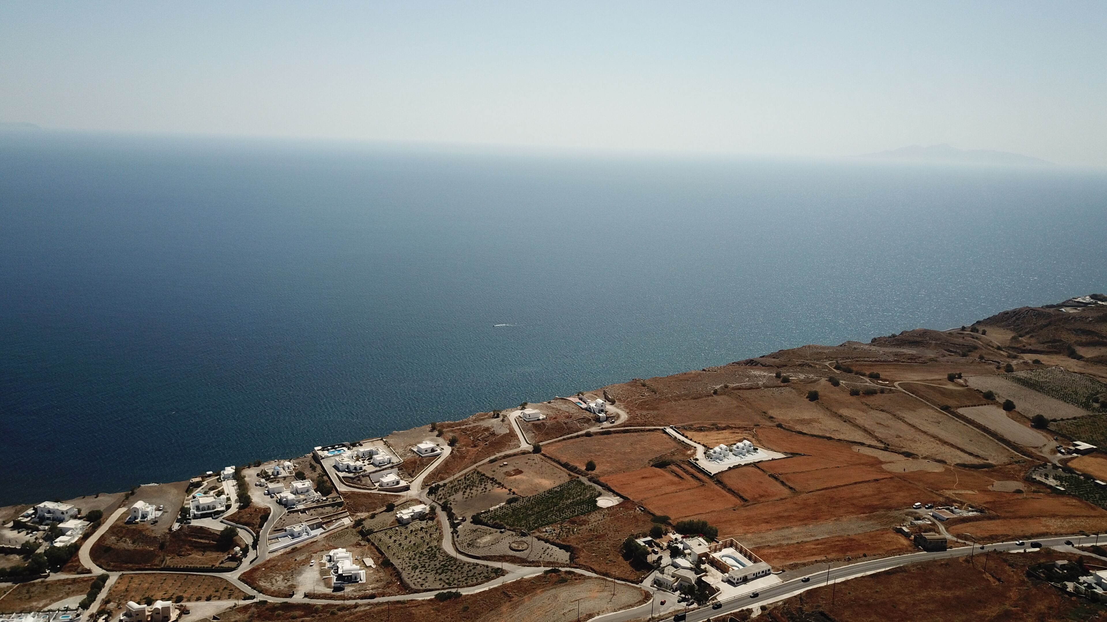 An aerial view of a coastal village with white buildings, terraced fields, and a road leading to the sea.