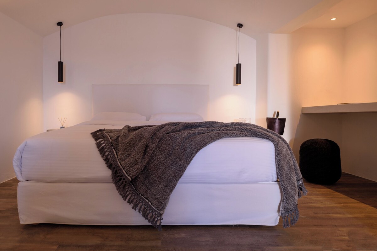 A modern bedroom with a large bed, two pendant lights, a wooden floor, and a small black ottoman.