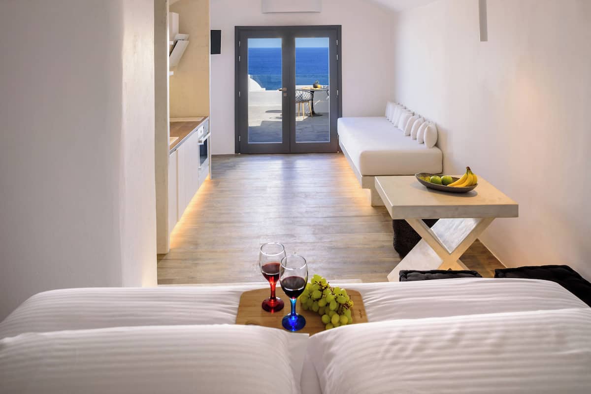 A hotel room with a bed, a dining table with a bowl of fruit and wine glasses, and a view of the ocean through a glass door.