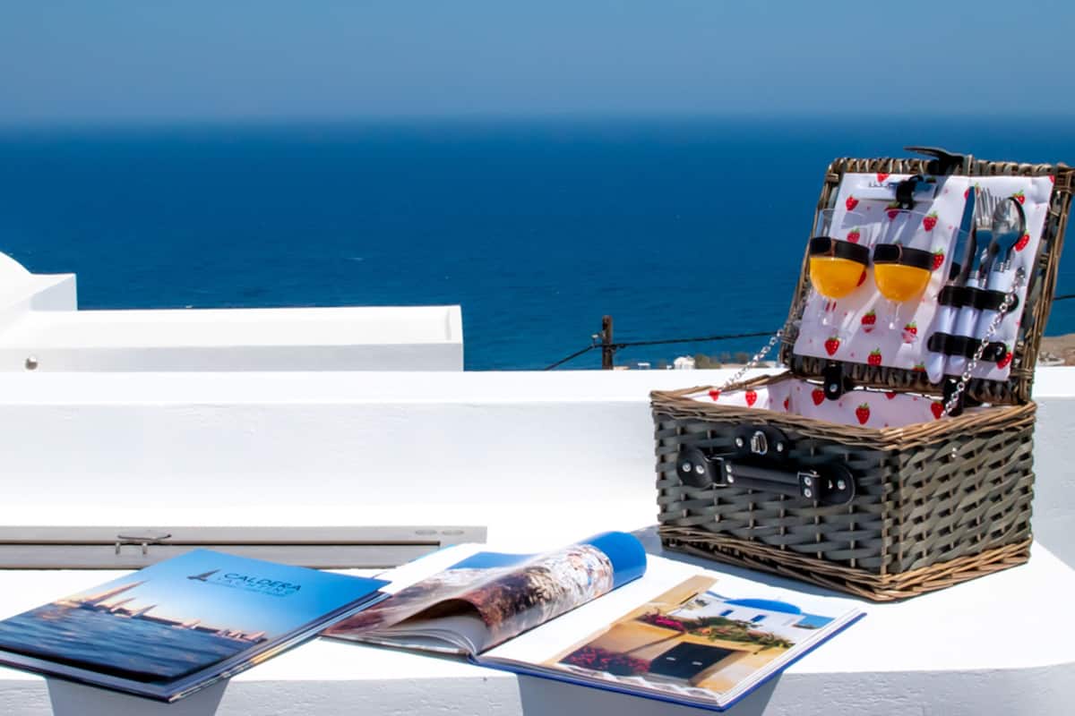 A wicker basket with a white cloth and sunglasses, a magazine, and a rolled-up brochure on a white surface with a view of the sea.