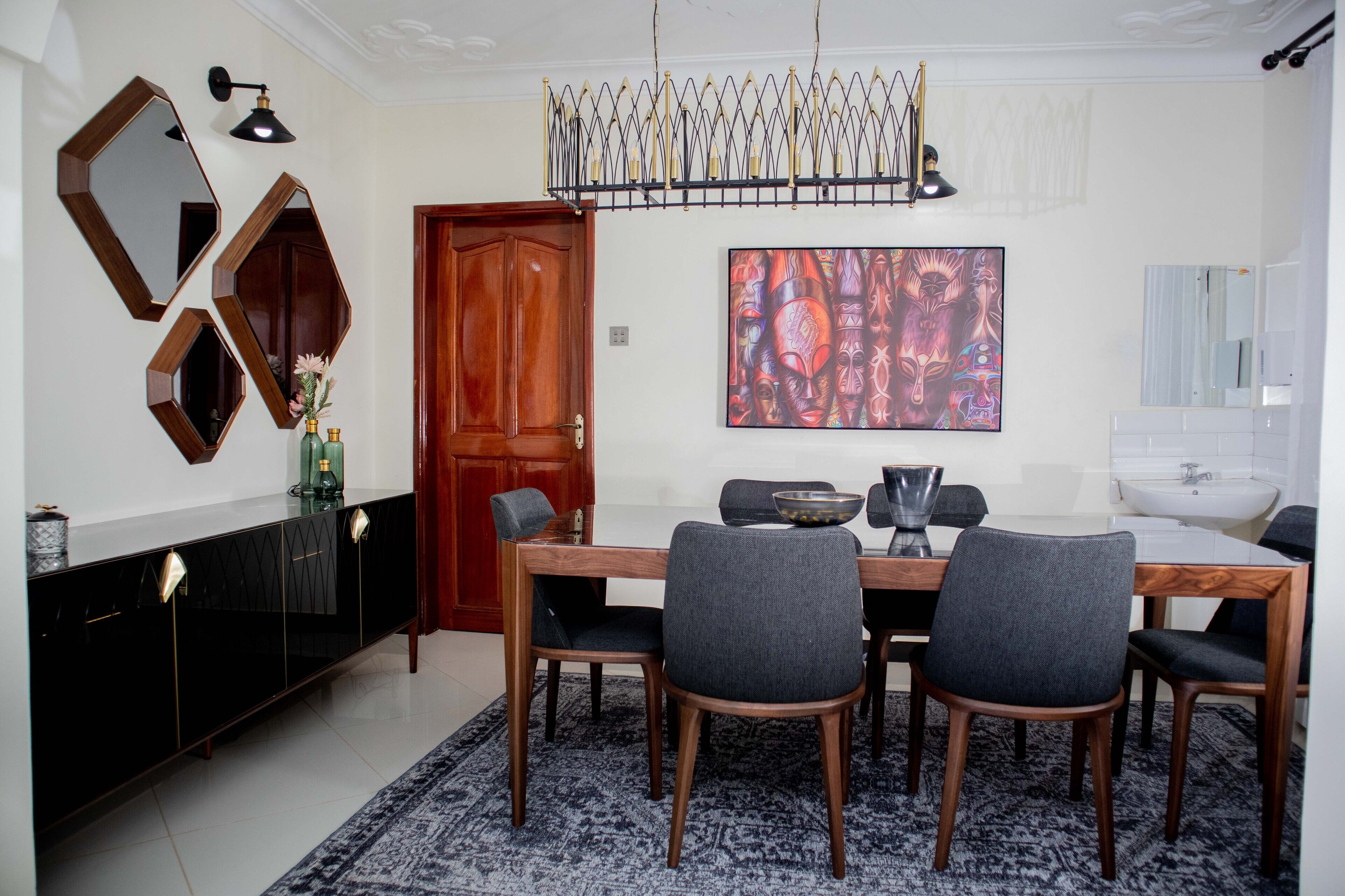 Un comedor con una mesa de madera, seis sillas grises, una consola negra con detalles dorados y un cuadro abstracto grande en la pared.
