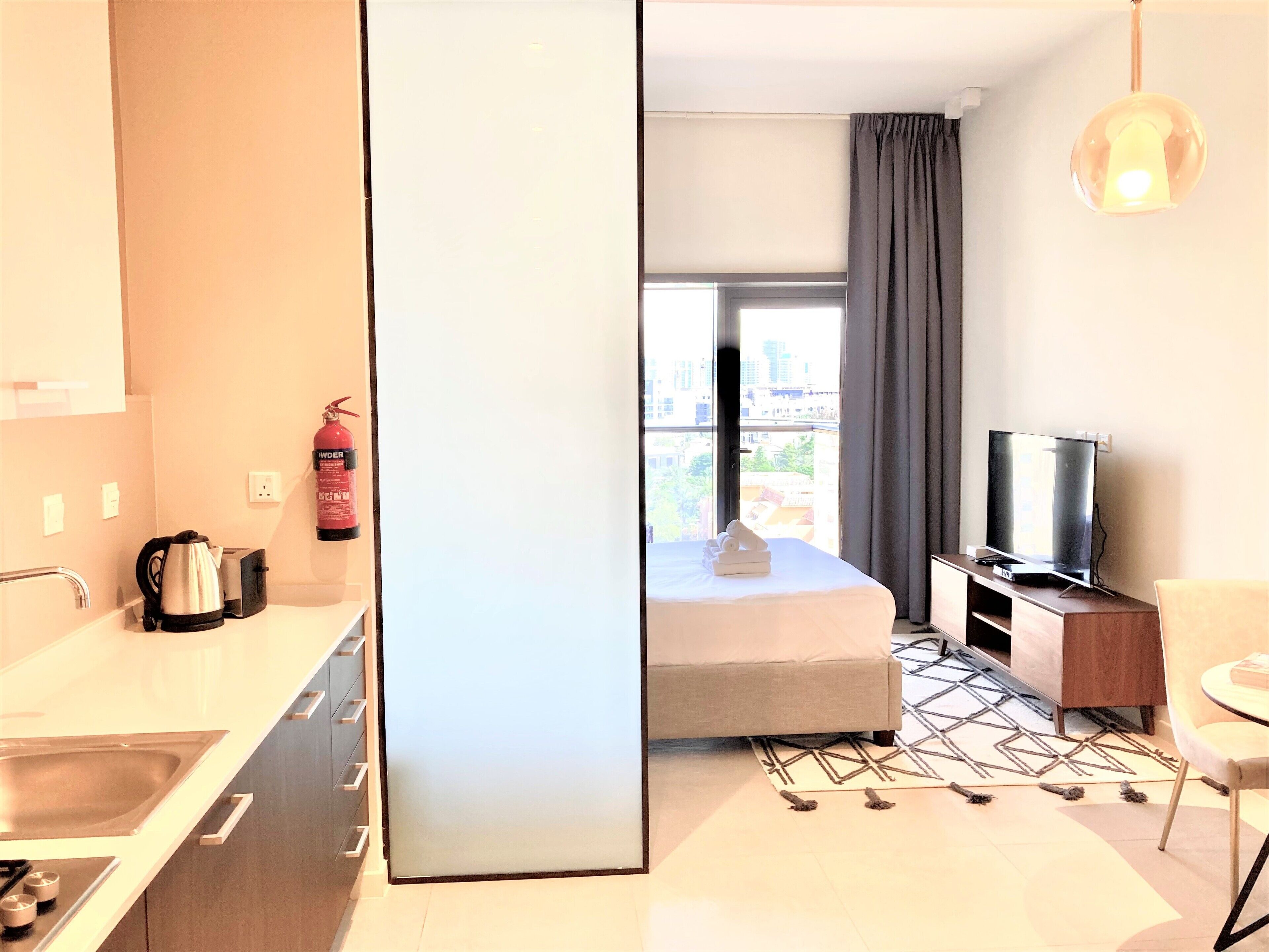 A hotel room with a kitchenette, a bed, a TV, and a view of the city.
