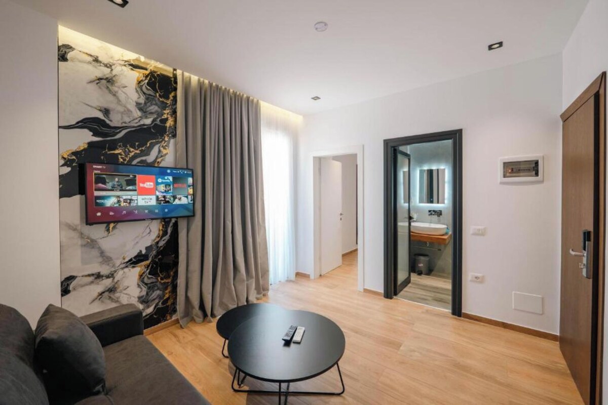 A modern living room with a flat-screen TV, a marble accent wall, a sofa, a round table, and a hallway leading to a bathroom.