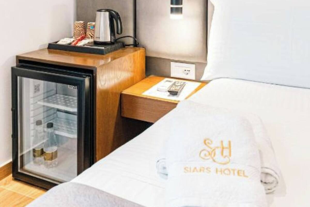 A hotel room with a bed, a nightstand with a lamp, a minibar, and a towel with the hotel logo.
