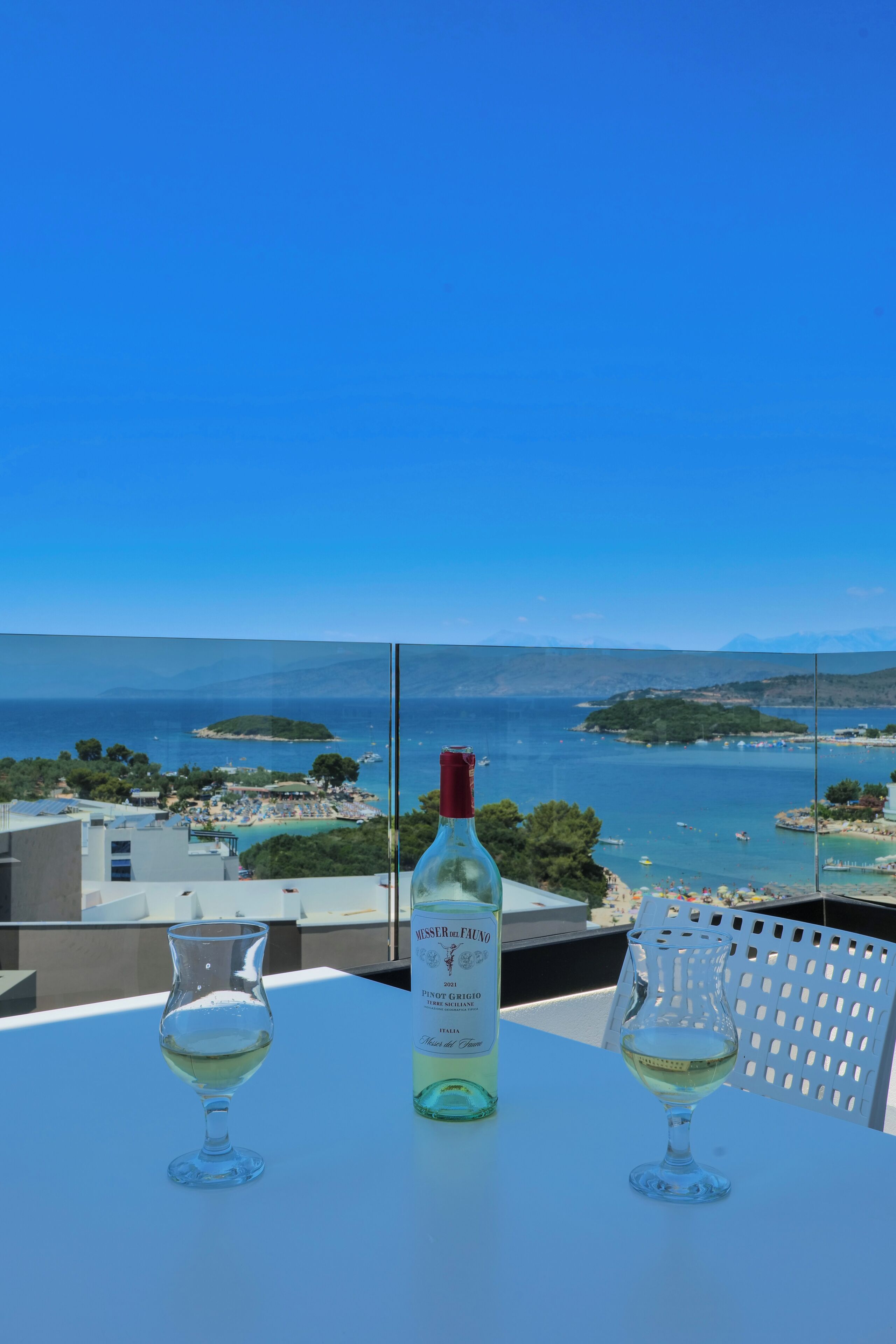A table with a bottle of white wine and two wine glasses on a balcony overlooking a coastal view.