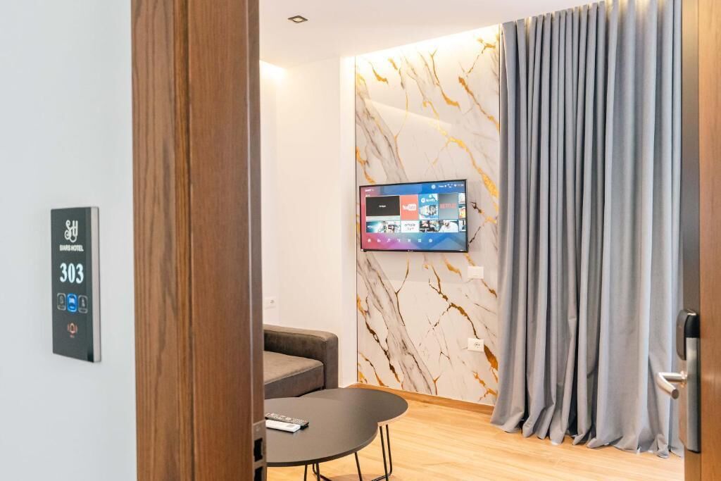 A hotel room with a marble-patterned wall, a mounted TV, a grey curtain, a wooden door, and a small table with a remote control.