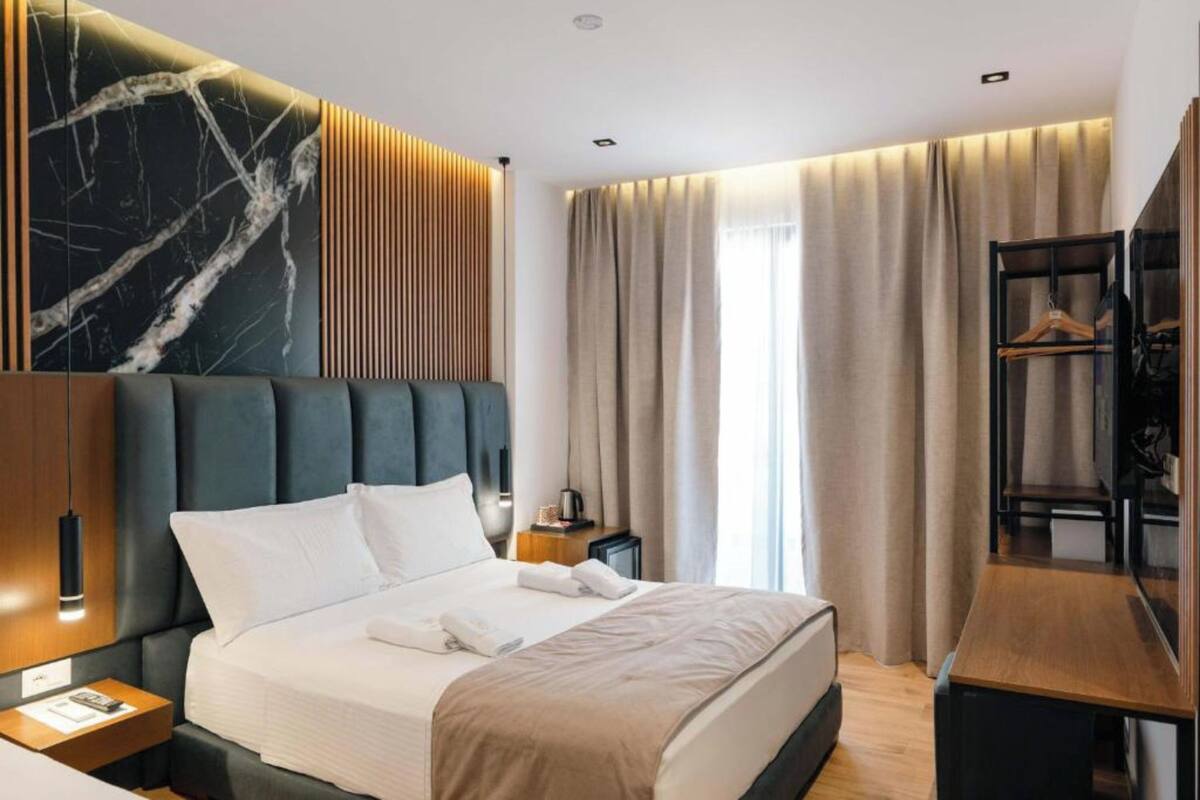 A modern hotel room with a large bed, a wooden nightstand, a desk, and a wardrobe.