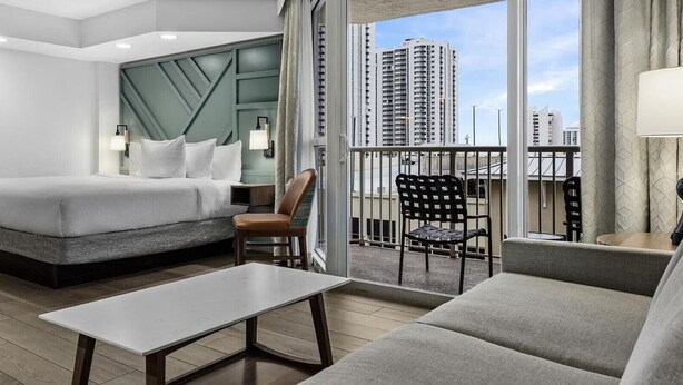 Una habitación de hotel moderna con una cama grande, un balcón con vistas a edificios altos, una mesita y una silla.