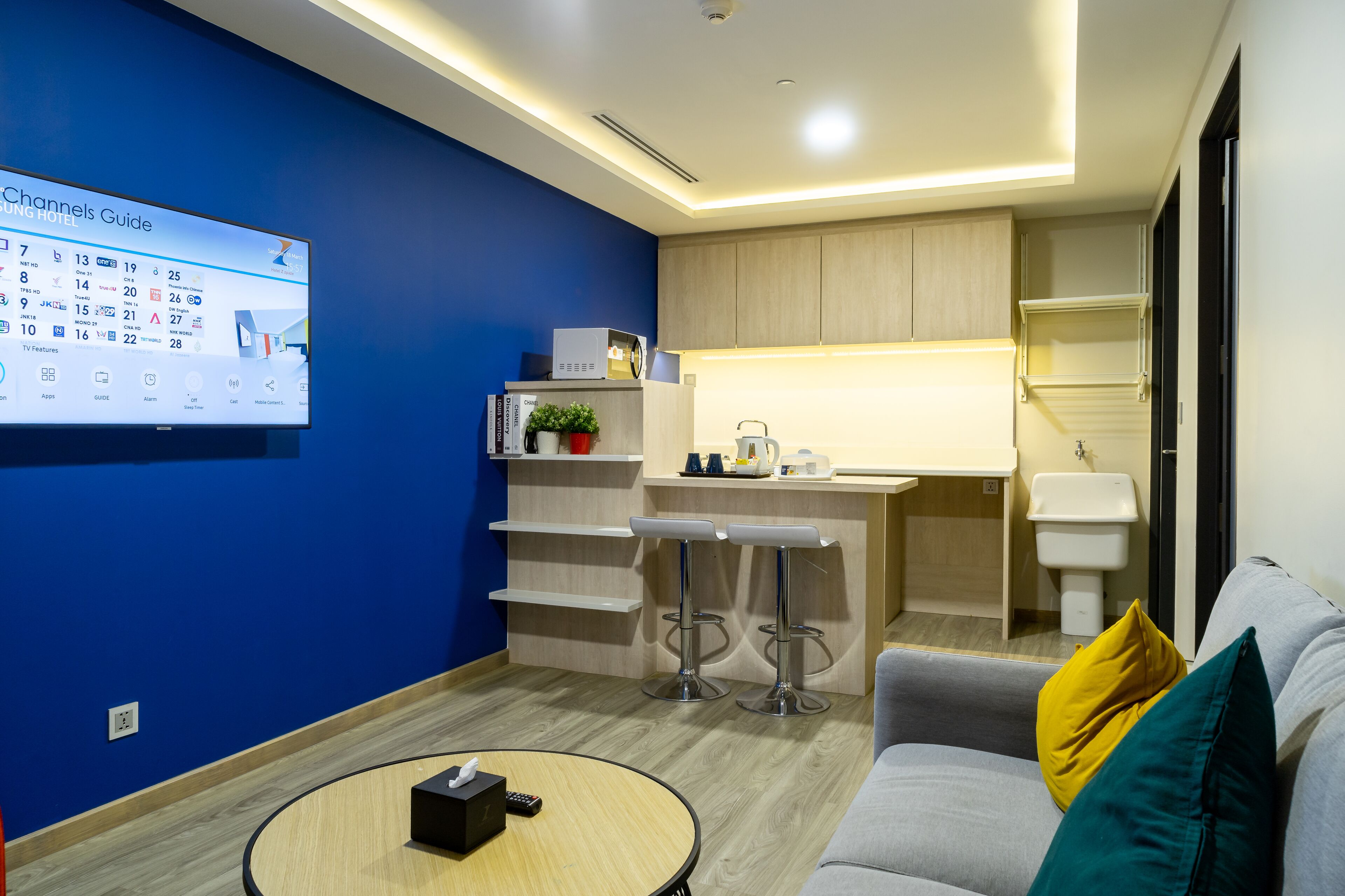A modern hotel room with a blue accent wall, a kitchenette, a bar area, and a seating area with a sofa and a coffee table.