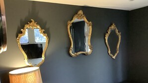 Een kamer met een donkere muur, drie versierde spiegels, een lamp met een gemêleerd lampscherm en een zwarte leren bank met decoratieve kussens.