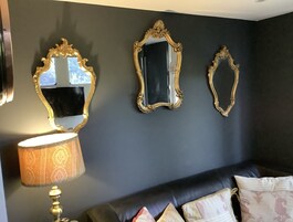 A room with a dark wall, three ornate mirrors, a lamp with a patterned shade, and a black leather sofa with decorative pillows.