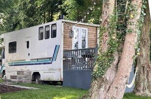 A white RV with a small attached wooden structure, possibly a shed or storage unit, parked next to a tree.