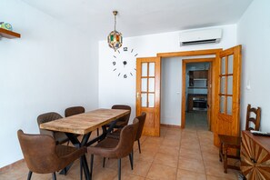 A dining area with a wooden table and chairs, a wall clock, and an open door leading to a kitchen.