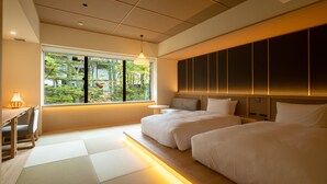 A hotel room with two beds, a small table, a chair, and a window with a view of trees.