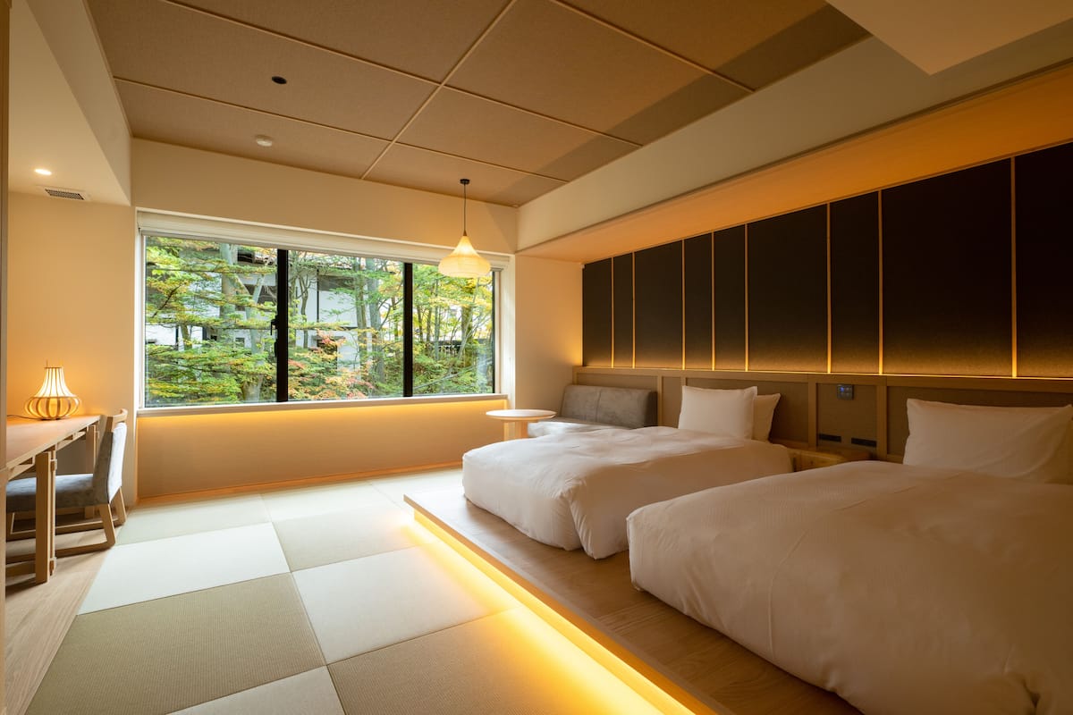 Ein Hotelzimmer mit zwei Betten, einem kleinen Tisch, einem Stuhl und einem Fenster mit Blick auf Bäume.