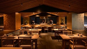 A restaurant with a high ceiling and wooden beams, featuring a central counter where chefs are preparing food. Tables and chairs are arranged for dining.