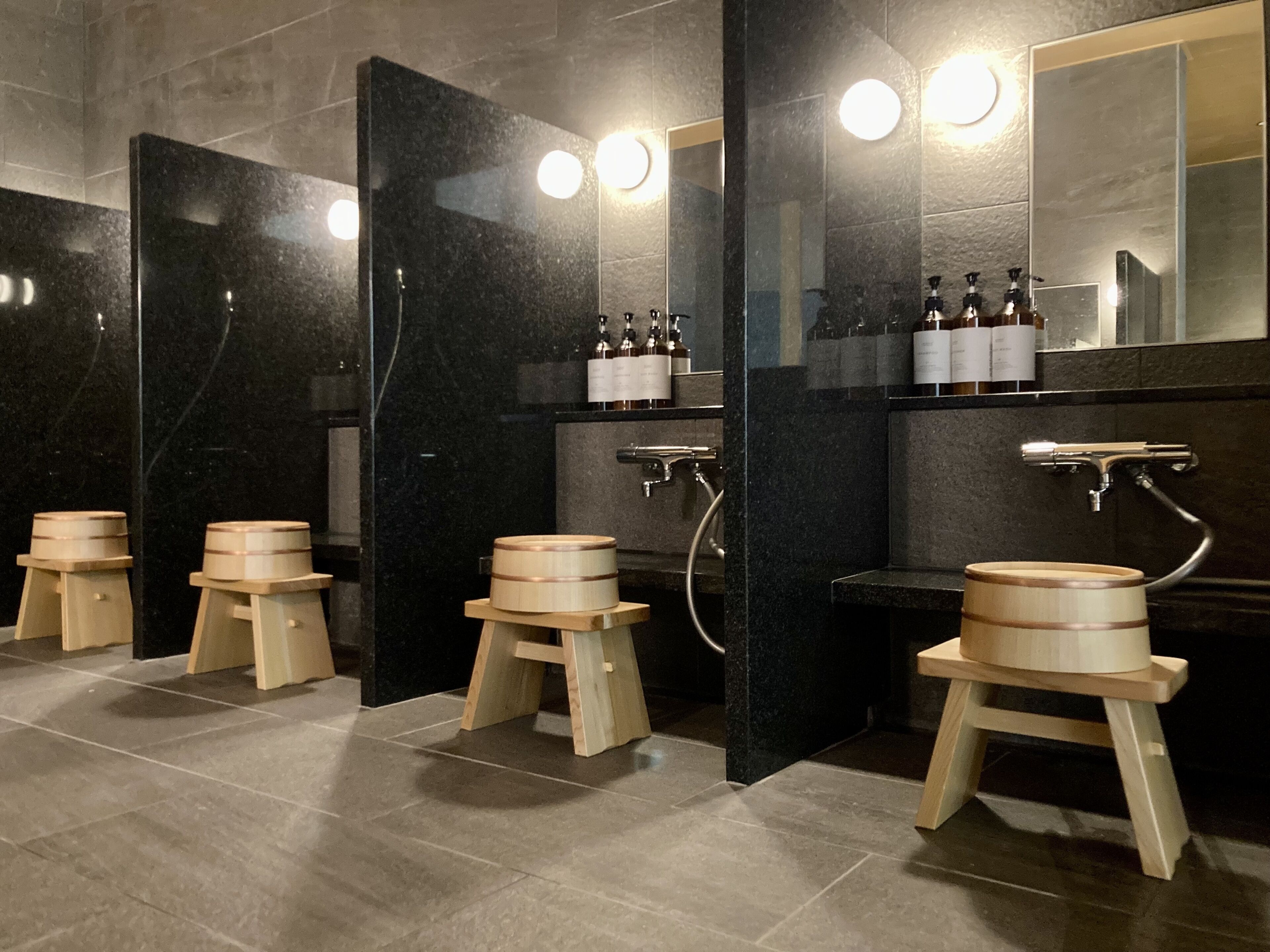 Un baño moderno con paredes de mármol negro, taburetes de madera y un espejo grande.
