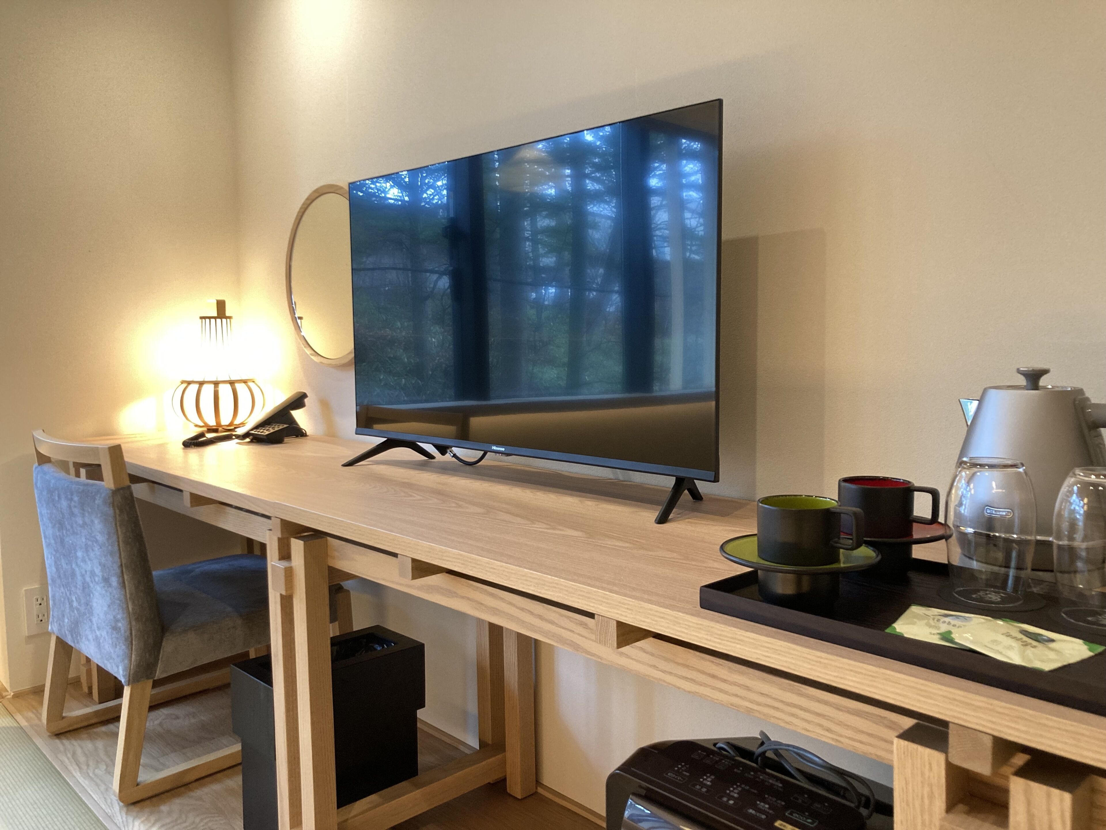 A modern room with a wooden desk, a flat-screen TV, a chair, a lamp, a coffee maker, and a trash bin.