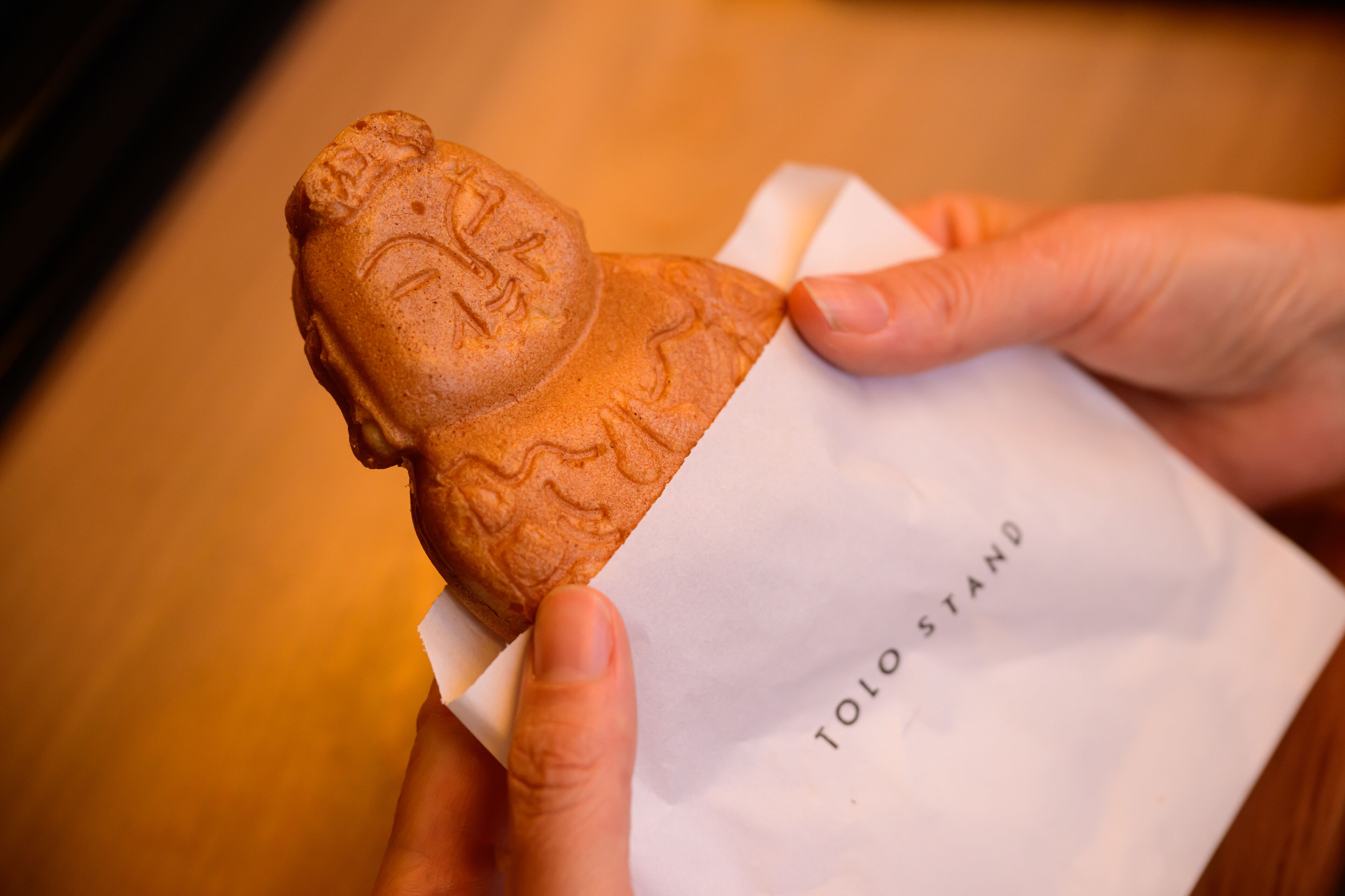Une main tenant un produit de boulangerie sur lequel est imprimé un visage, posé sur une feuille de papier blanche portant l’inscription « TOLO STAND ».