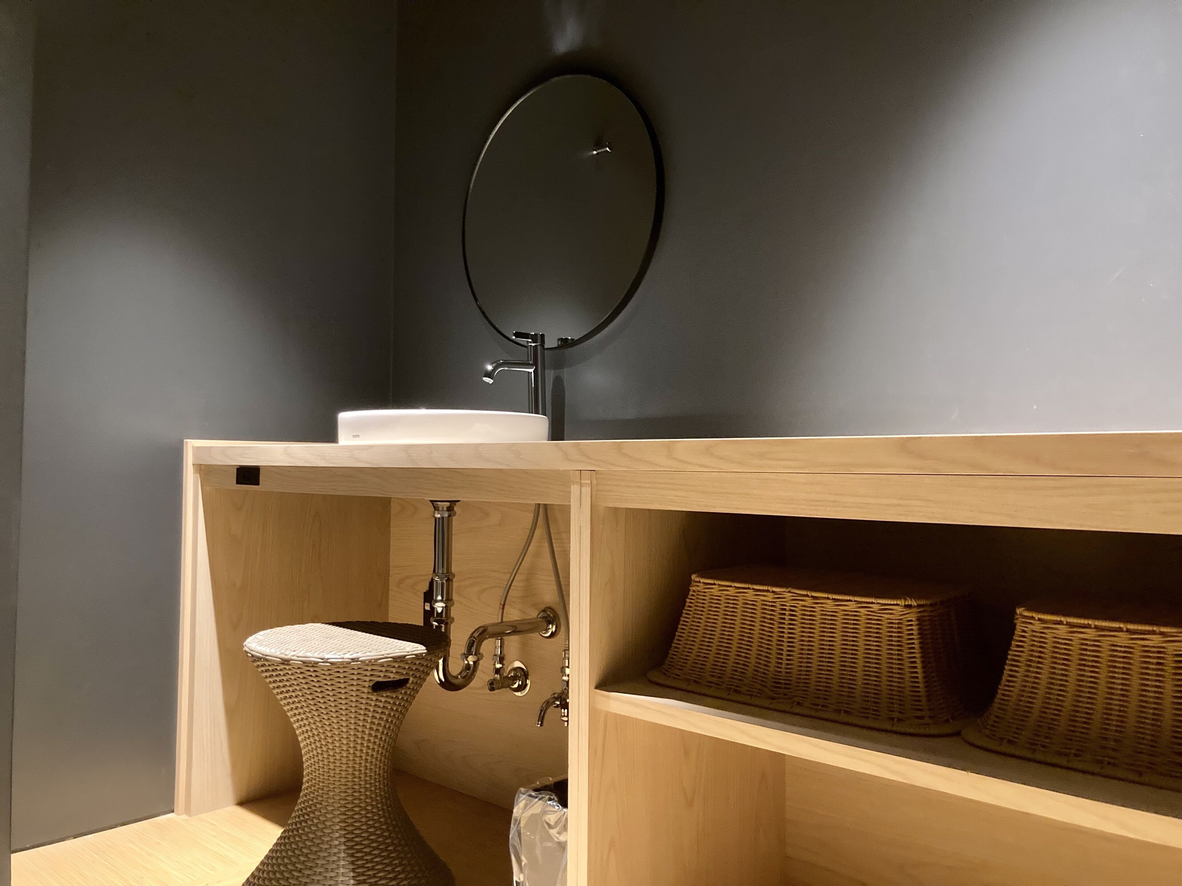 A bathroom with a dark wall, a round mirror, a white sink, a wicker vase, and wicker baskets on shelves.