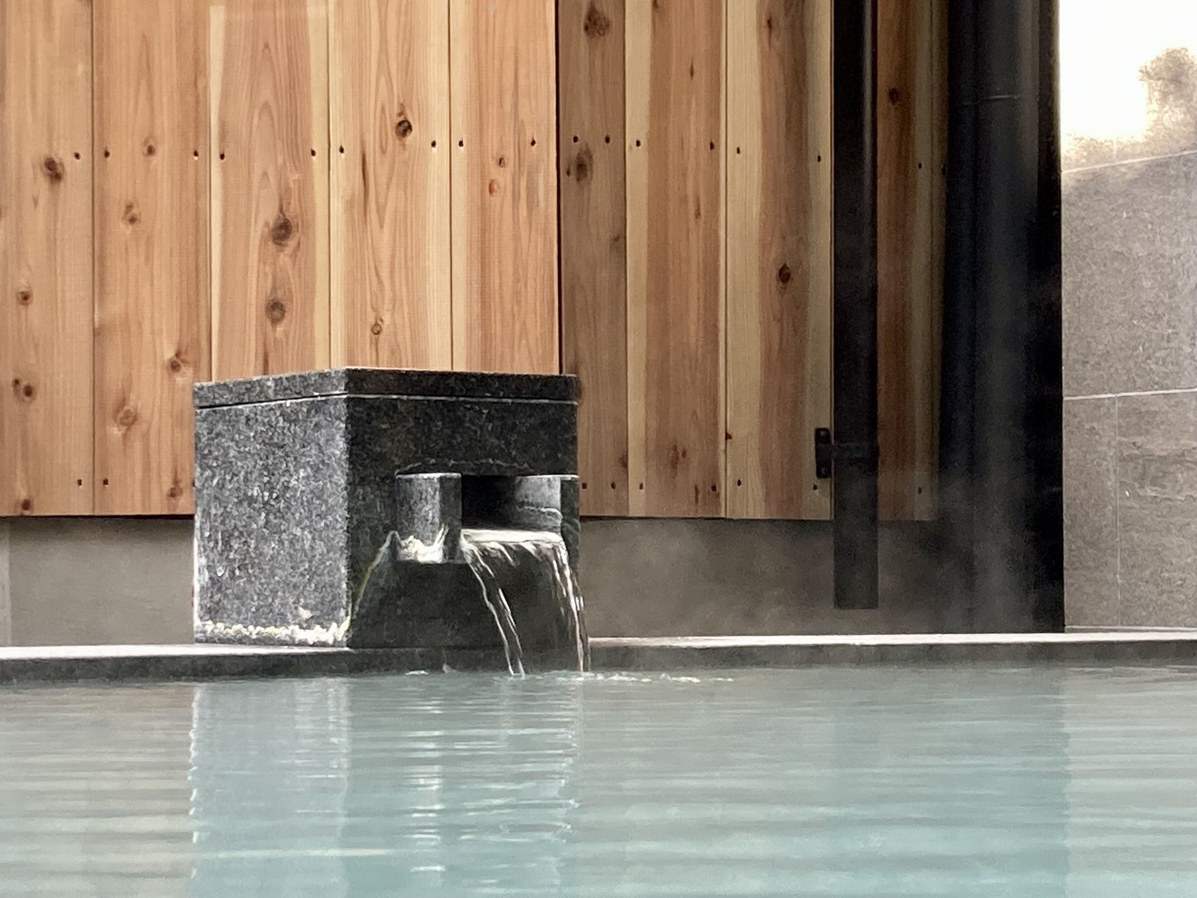 A stone hot tub with a waterfall feature in front of a wooden wall.