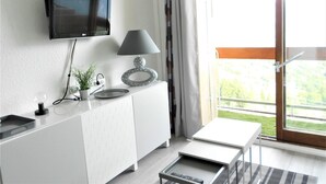 A modern living room with a flat-screen TV mounted on the wall, a white cabinet with a hexagonal tile backsplash, a small table with a lamp, and a sliding glass door leading to a balcony with a view of greenery.