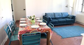 Uma sala de jantar com uma mesa posta para quatro pessoas, cadeiras, um sofá e um abajur.
