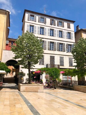 Een binnenplaats met een gebouw met meerdere ramen en luiken, omgeven door bomen en potplanten.
