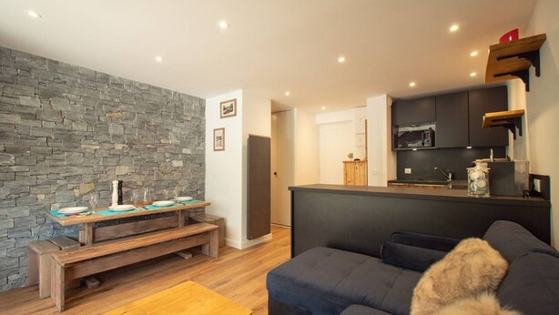 Un salon moderne avec un mur en pierre, un canapé foncé, une table basse en bois et un coin cuisine doté de meubles sombres et d’un four à micro-ondes.