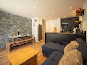 Een moderne woonkamer met een stenen muur, een donkere bank, een houten salontafel en een keukenhoek met donkere kastjes en een magnetron.