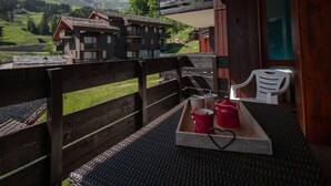 Un balcon doté d’une balustrade en bois, une table sur laquelle se trouve un plateau avec des tasses rouges et un dessous de verre en forme de cœur, et une vue sur un paysage montagneux avec des bâtiments en arrière-plan.