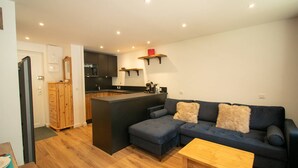 Un espace de vie moderne avec un canapé bleu foncé, une table basse en bois et un coin cuisine doté de meubles de rangement sombres et d’une armoire en bois.