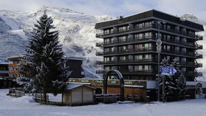 Um hotel com vários andares e varandas, com uma estância de esqui ao fundo e uma placa indicando um teleférico.