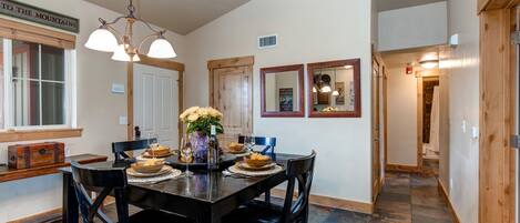 A dining area with a wooden table, black chairs, and a chandelier. There are framed pictures on the wall and a window with a view of the mountains.