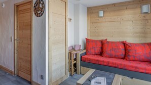 A room with a wooden door, a red sofa with patterned cushions, a small table with a remote control and newspaper, and a wall clock. - Renovated Studio 2 pers · Wellness · Pets allowed (La Plagne Montchavin Les Coches)