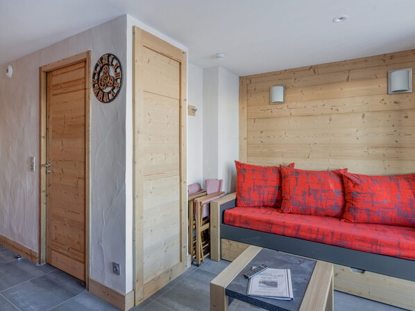 A room with a wooden door, a red sofa with patterned cushions, a small table with a remote control and newspaper, and a wall clock. - Renovated Studio 2 pers · Wellness · Pets allowed (La Plagne Montchavin Les Coches)