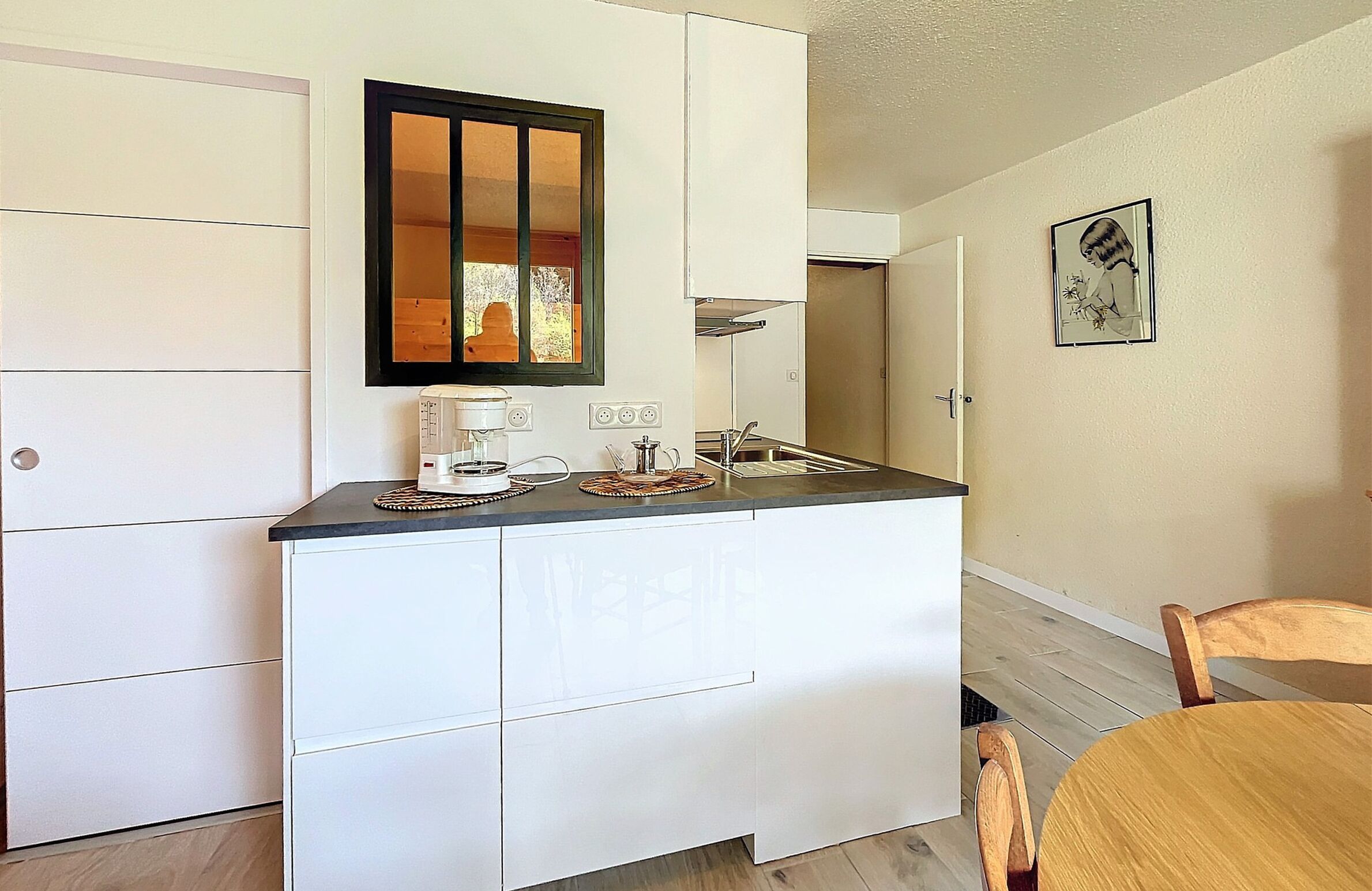 A compact kitchen with a built-in stove, a sink, and a small countertop. There is a wooden dining table with chairs, and a framed picture on the wall.