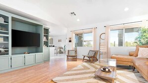 A spacious living room with a large flat-screen TV, a wooden coffee table, and a beige rug. There is a sofa and two wicker chairs.