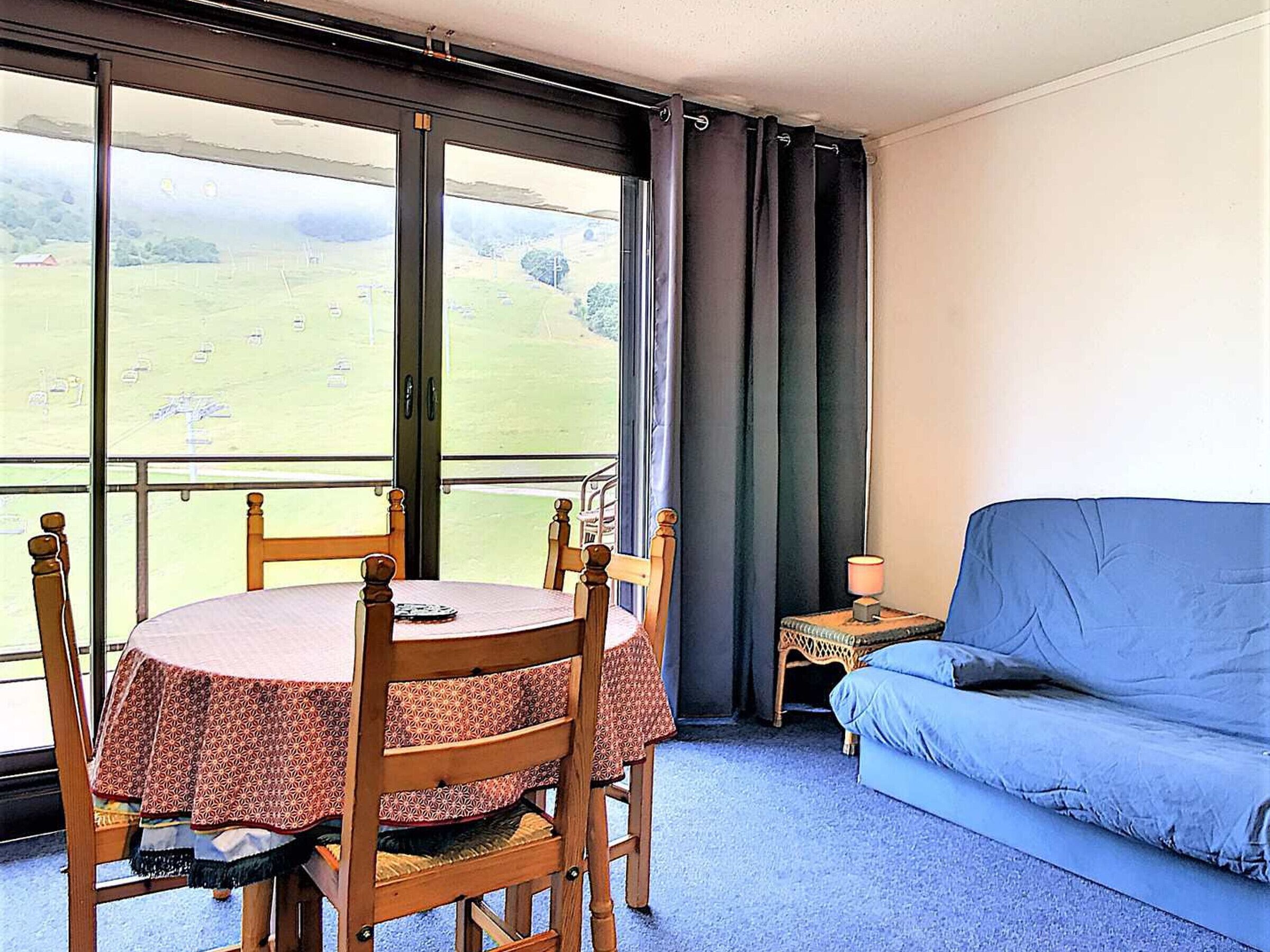 A room with a balcony, a round table with chairs, a blue sofa, and a small table with a lamp.