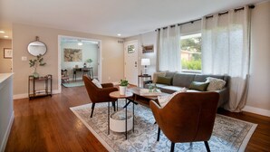 Una sala moderna con un sofá, dos sillones, una mesa de centro y una zona de comedor con mesa y sillas. Hay un ventanal con cortinas, un ventilador de techo y un espejo en la pared.
