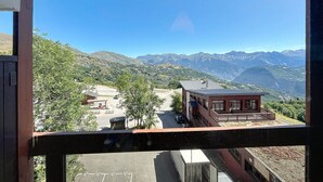 Vista de uma varanda com paisagem montanhosa, com um edifício que também tem varanda e uma área pavimentada com um carro e uma caminhonete.