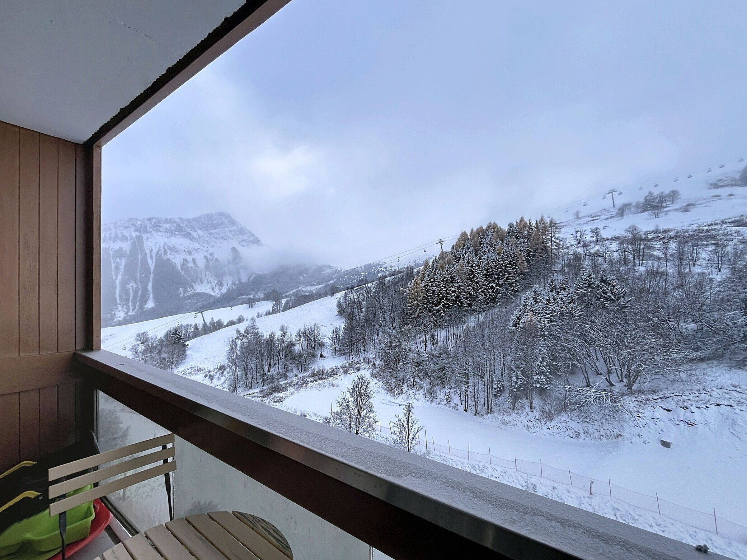 Vue depuis un balcon doté d’une balustrade en bois, donnant sur un paysage montagneux enneigé, avec des arbres et un télésiège visible au loin.