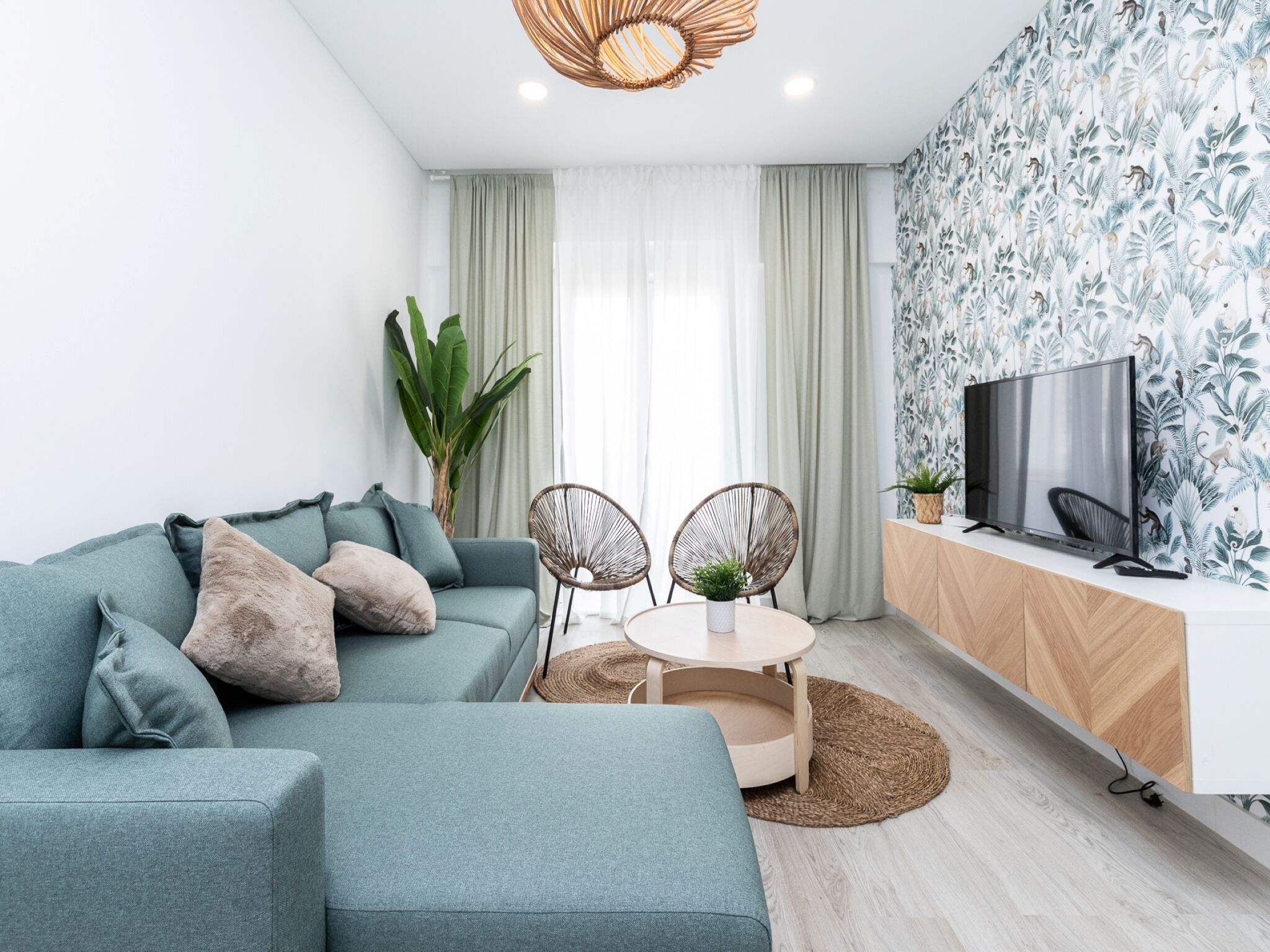 A modern living room with a sectional sofa, a round table with two chairs, a television on a wooden cabinet, and a large window with sheer curtains.
